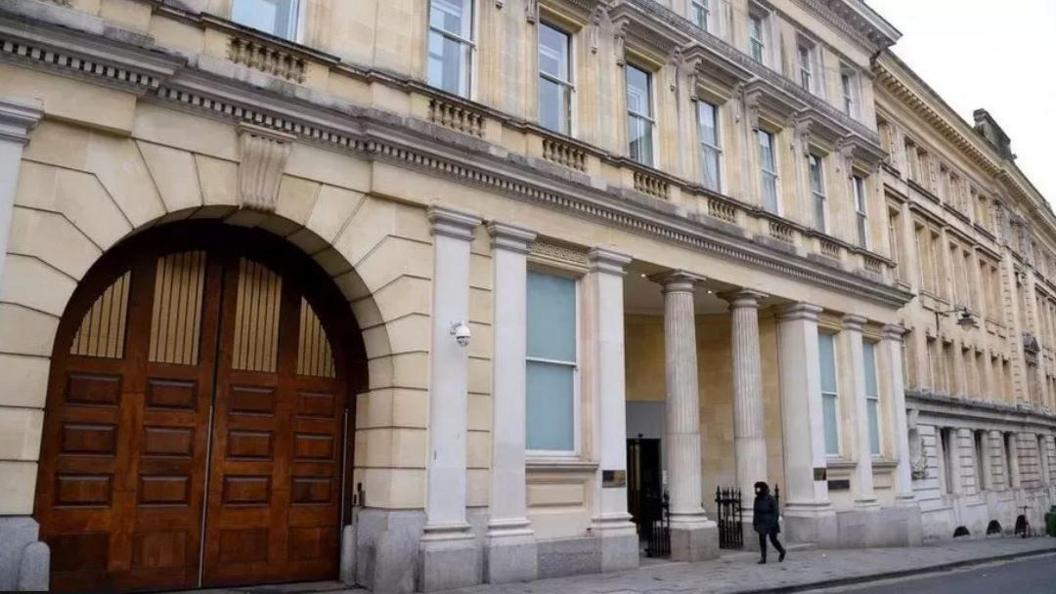 Bristol Crown Court as seen from the road outside. It is a large sandstone coloured building with pillars and a rounded wooden gate on the left. 