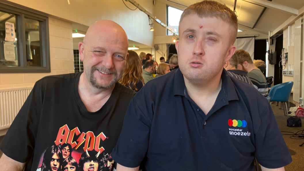 Two men look at the camera. One wears a T-shirt with an ACDC logo on the front while the second is in a polo shirt with the words Worcester Snoezelen on it