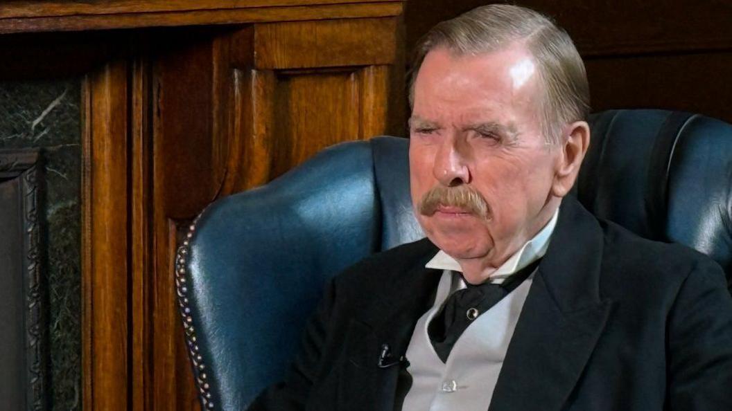 Actor Timothy Spall on the set of Night and Day. He is sitting in a blue chair and wearing a black suit jacket with a grey waistcoat and a black tie. He has a large moustache