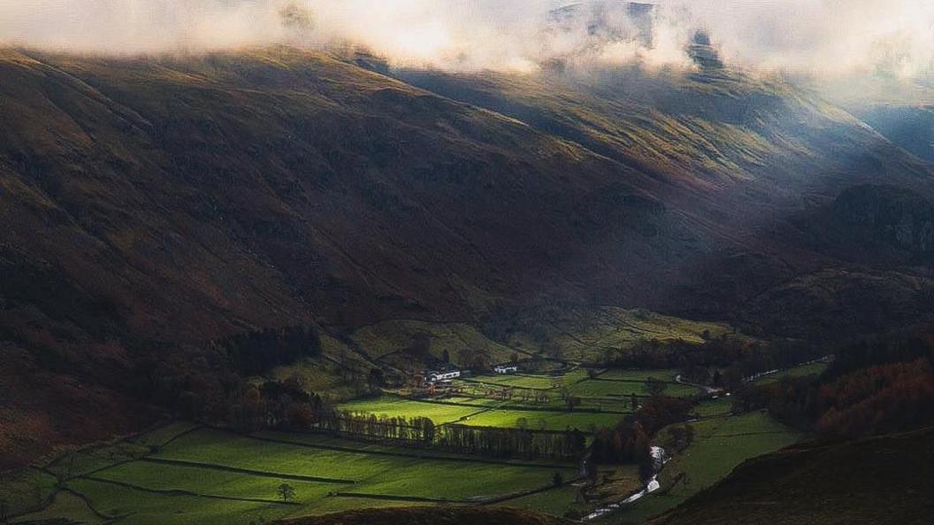 View from High Rigg. A sunbeam cuts across the valley and lights it up green. The rest of the landscape is shrouded in darkness and clouds.