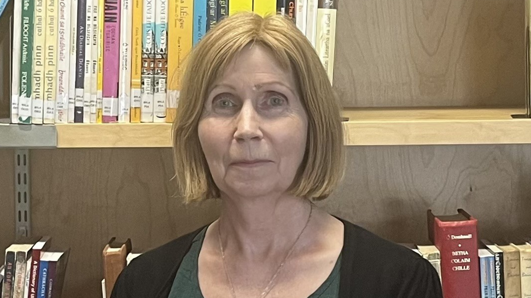 Linda Ervine, a woman with blonde hair styled in a bob, looks directly at the camera.  She is standingi front of a wooden bookshelf.  She is wearing a black cardigan over a dark green top. 