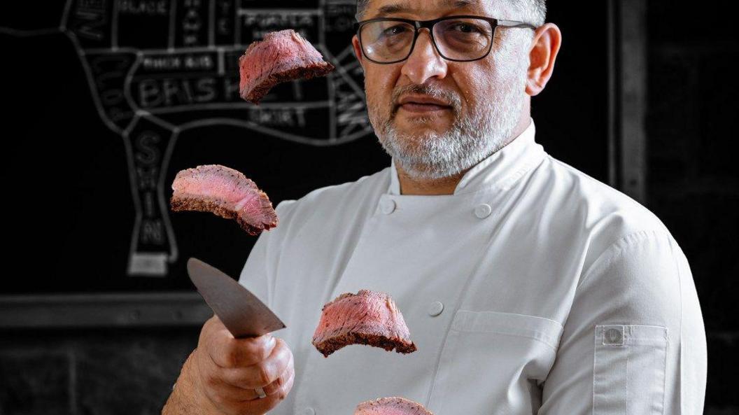 A man wearing a chef's white top stands holding a very sharp knife and looking at the camera while multiple pieces of steak are seen airborne in a picture taken using time delay technology. The man, who is a member of staff at the Ashville Steak House in Southville in Bristol, has a black and white beard and is wearing glasses