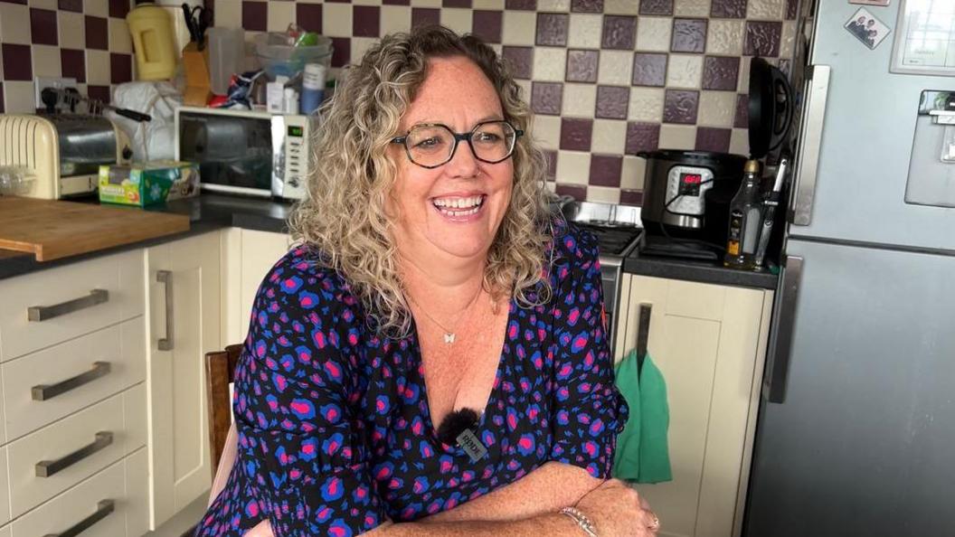 Liz Ball is sitting in a kitchen. She has curly hair and is smiling 