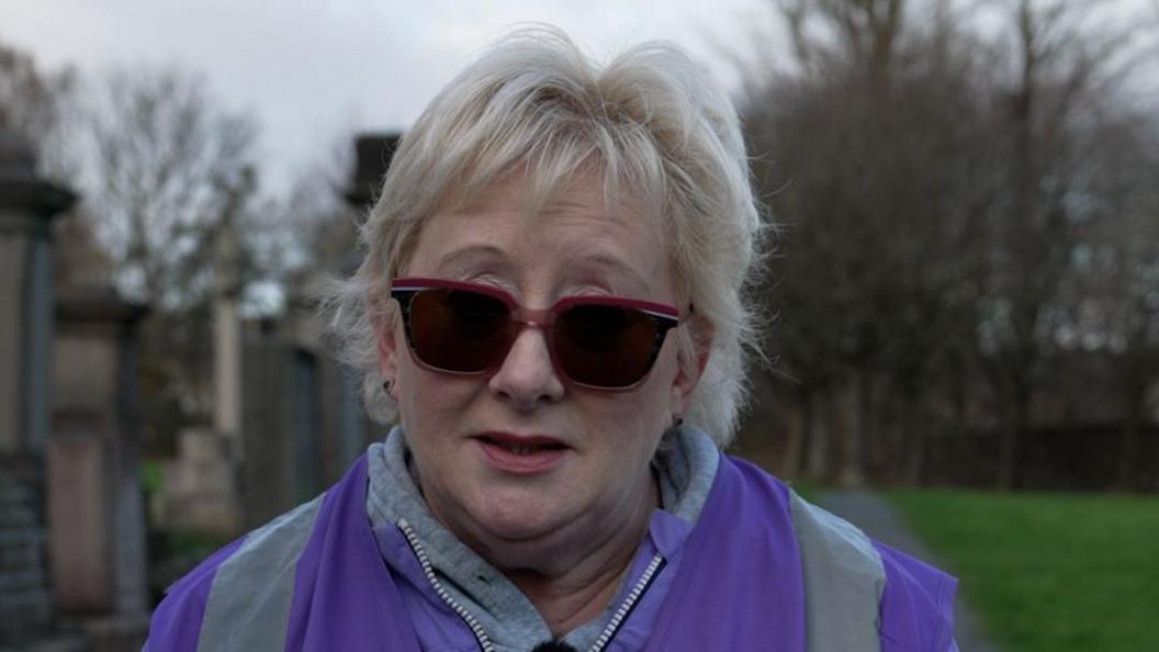 Annette Mullen, who has blonde hair and sunglasses, is wearing a purple high vis jacket and standing in a cemetery with a row of graves behind her. 