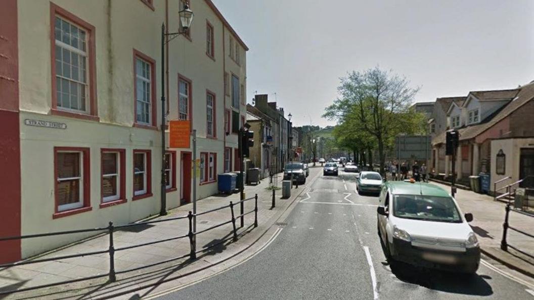 Strand Street in Whitehaven. A white car drives towards the camera. A white building with red painted window frames stands on the right. The street sign is attached to the building. A row of trees can be seen lining the road further down the street on the right.