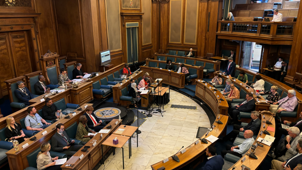 The main chamber inside Nottingham's Council House. People are sat around a semi circle table with straight rows in front.