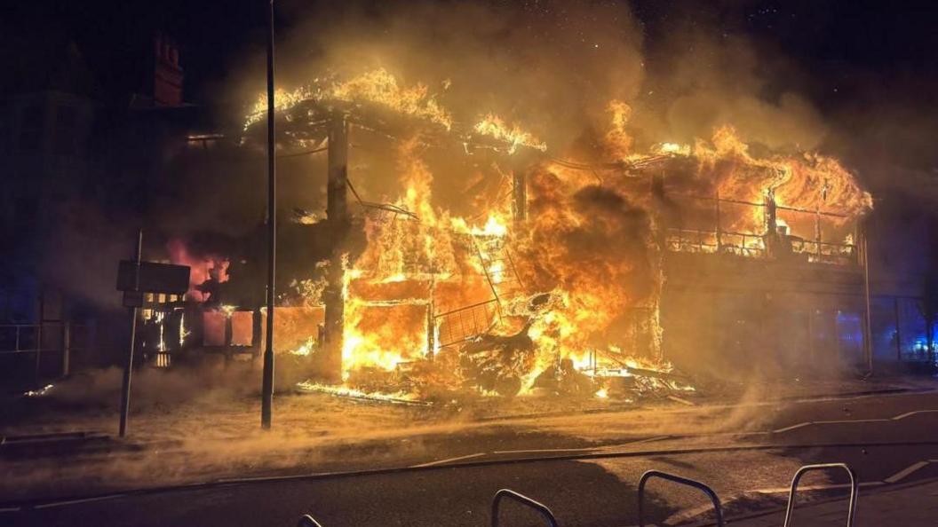 A building that is completely alight. Huge flames lick the frame of the building and there is a lot of smoke. It is night time.