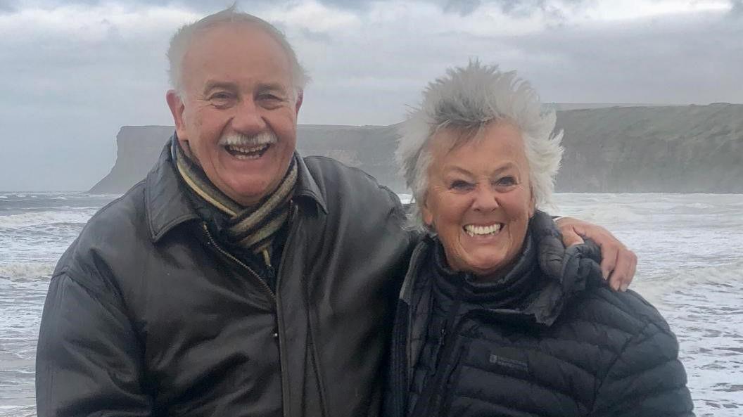 A smiling couple in their 70s stands in front of a seafront vista, with coastal and mountainous terrain in the distant background. The man is on the left, with grey hair and a moustache and dressed in a leather jacket, has arm around a woman with silver windswept hair and a puffer jacket on the right. 