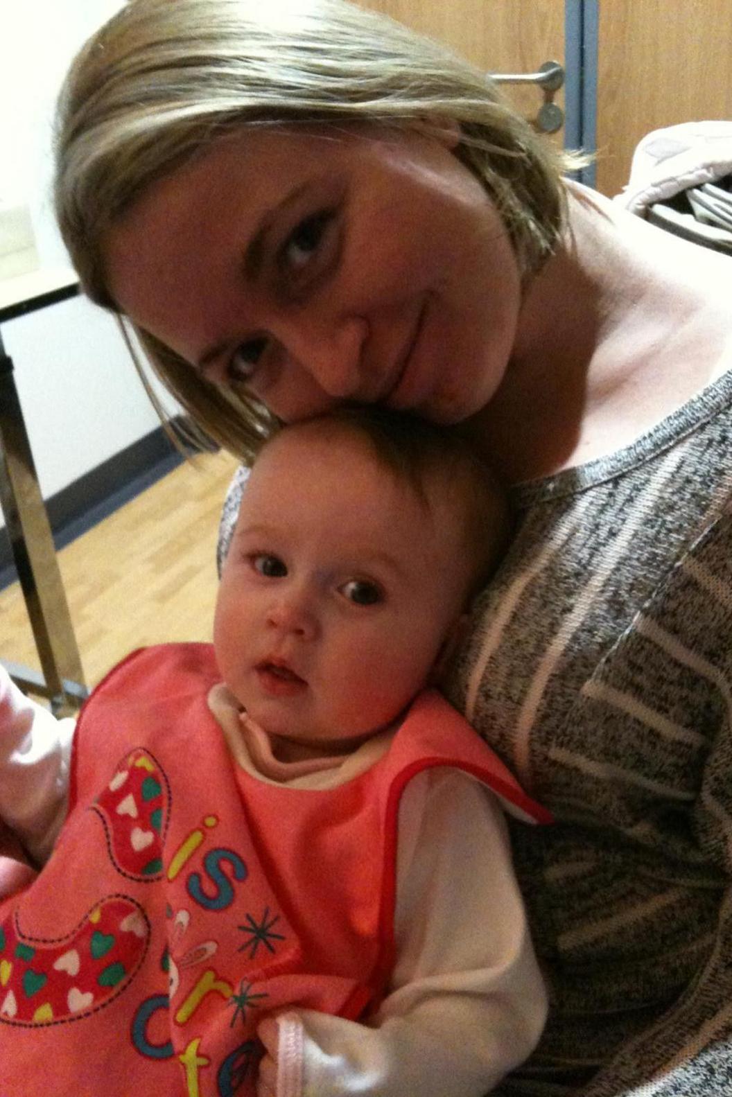 A woman in her early thirties with a short blonde bob and wearing a long sleeved grey t-shirt with white stripes and a scooped-neck, sits with a baby on her lap. She is smiling into the camera, while resting her cheek on top of the baby's head. The baby, which is a girl of around 8 months old, is also looking into the camera. She has some brown hair visible under the woman's chin and is wearing a pale pink top and a bright pink bib which is embroidered with hearts and letters. A pale wood door with a silver handle, and pale wooden flooring is visible in the background.