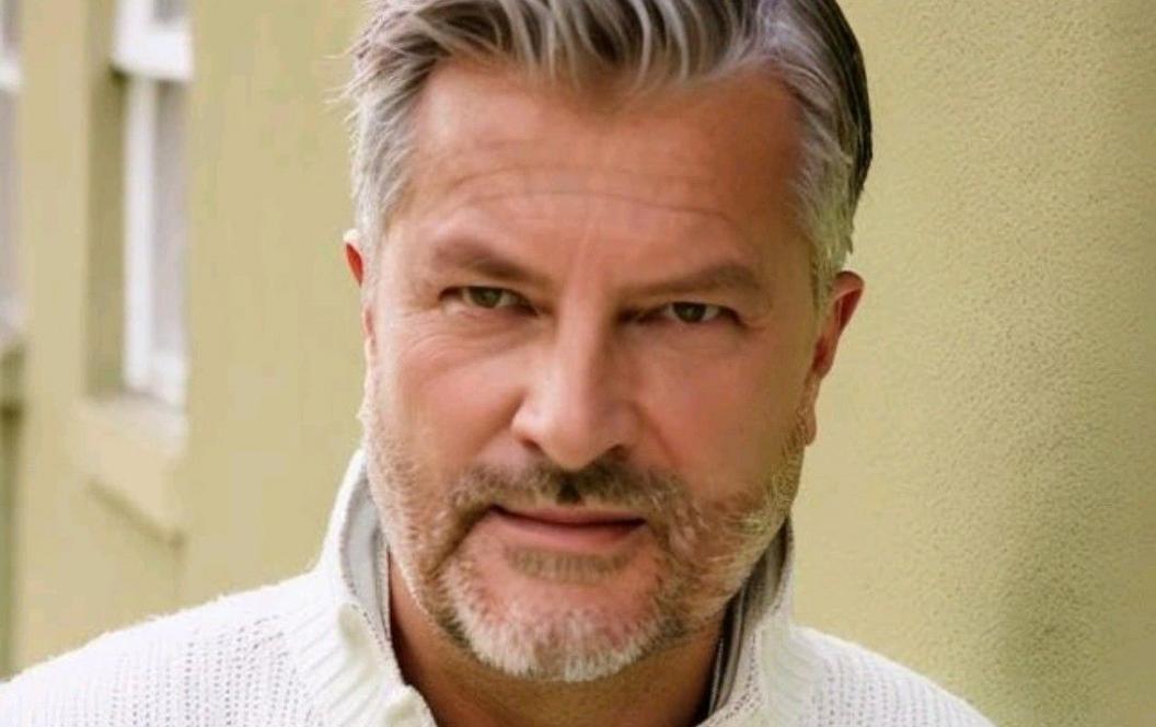 Simon Strutt, a man with short hair and a beard, wearing a white jumper, standing against a wall