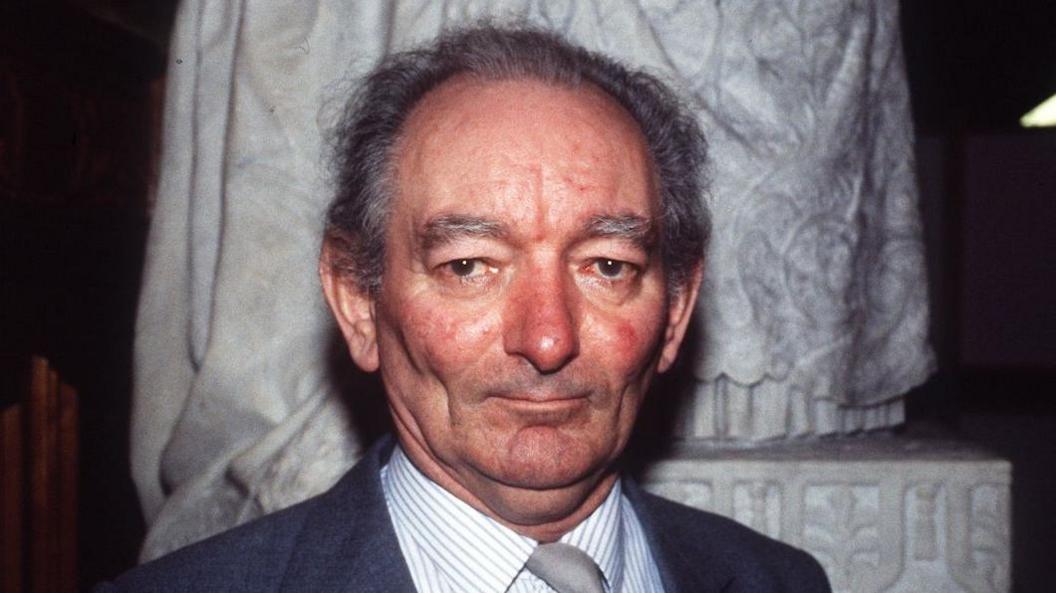 Brian Friel is looking directly at the camera, he has short wispy grey hair. He is wearing a suit and tie.