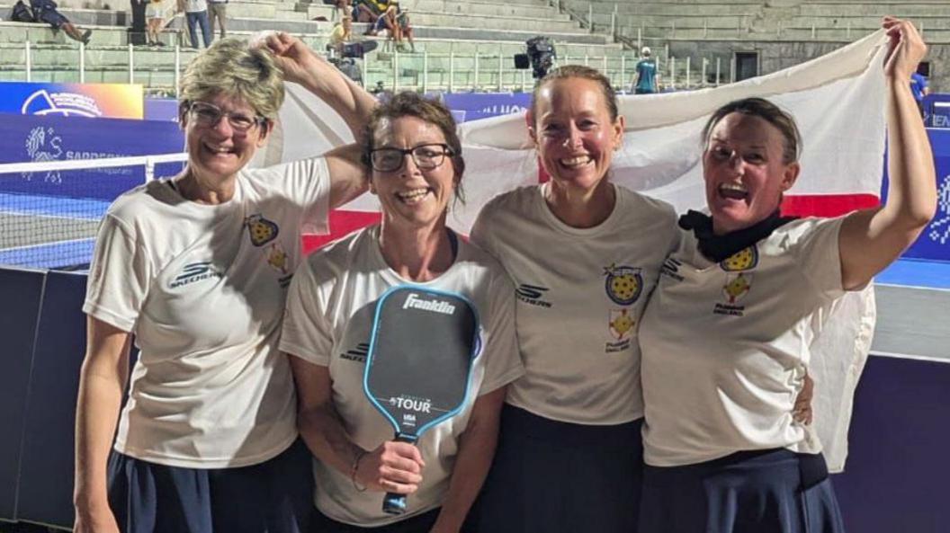 Four women, in white t-shirts and blue skirts. The two on the ends are holding up a English flag behind then. The one second to the left is holding a pickleball racket. All four are smiling. Behind them is the court. 