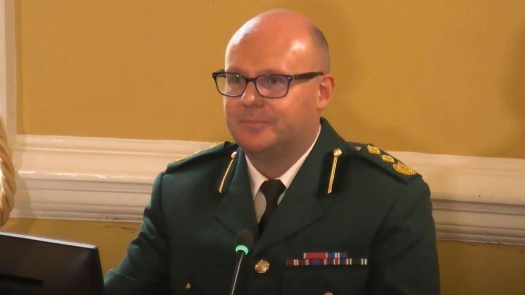 Mr Darch wearing a green military uniform with coloured patches. He is wearing dark framed glasses, has a bald head and is clean shaven. He is looking to the left of the camera at Mr Mansfield. 