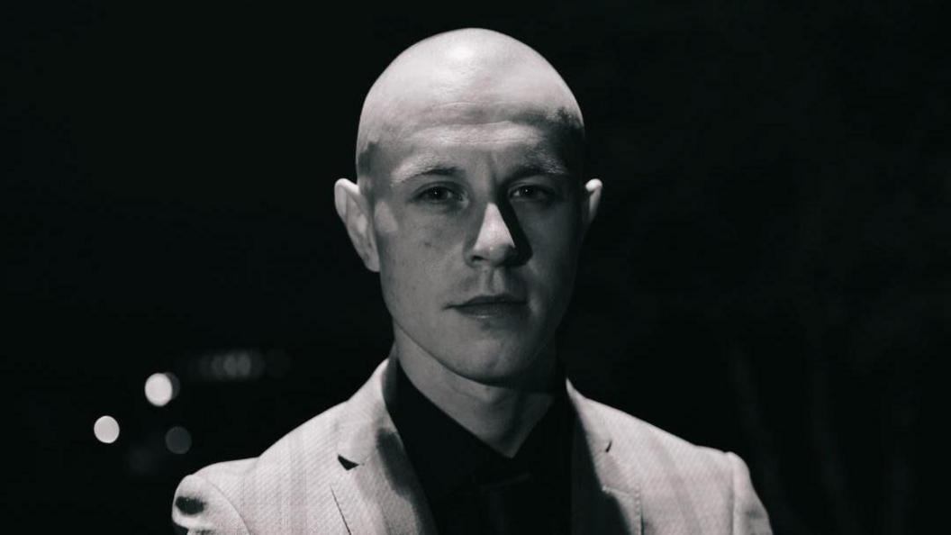 A black and white photo of a bald man. He wears a suit jacket and shirt. The shot is a close-up as he looks into the camera. 