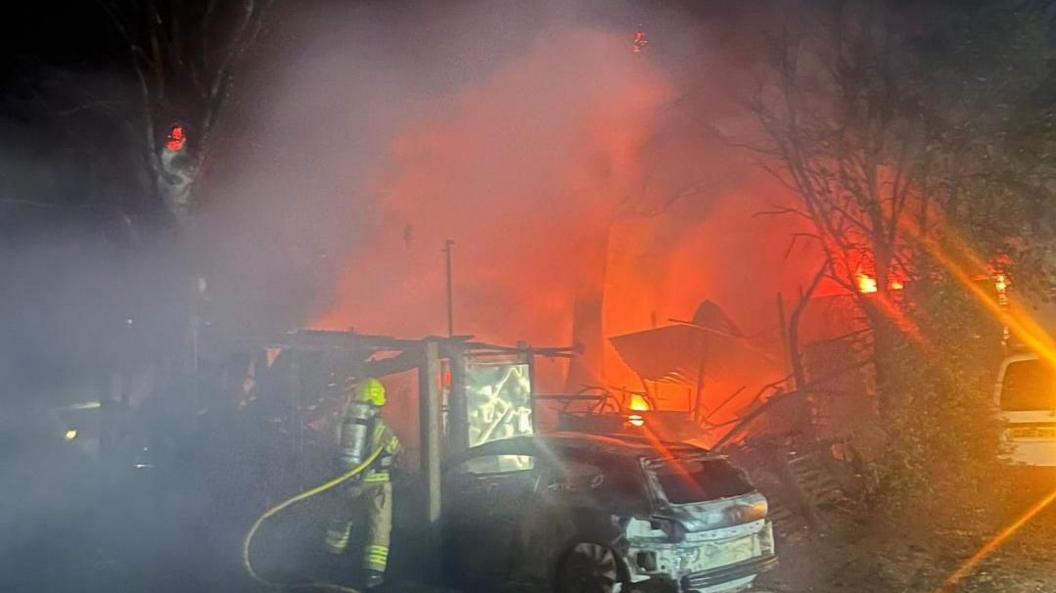The orange glow of a fire, and smoke billowing from it, can be seen in the background at night time. In the foreground is a firefighter, with his back to the camera and wearing the brown and yellow uniform. There is a damage car in the foreground too.