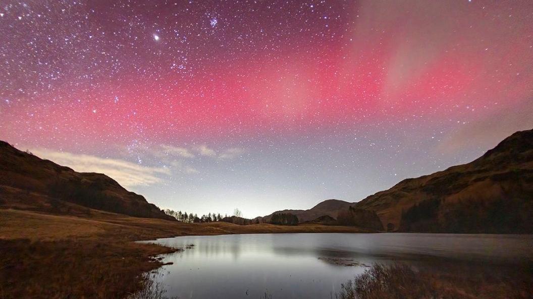 Northern Lights are seen in pink, red and purple, and lit up by stars over Blea Tarn in the Lake District. They are pictured over a lake with hills on either side of the valley.