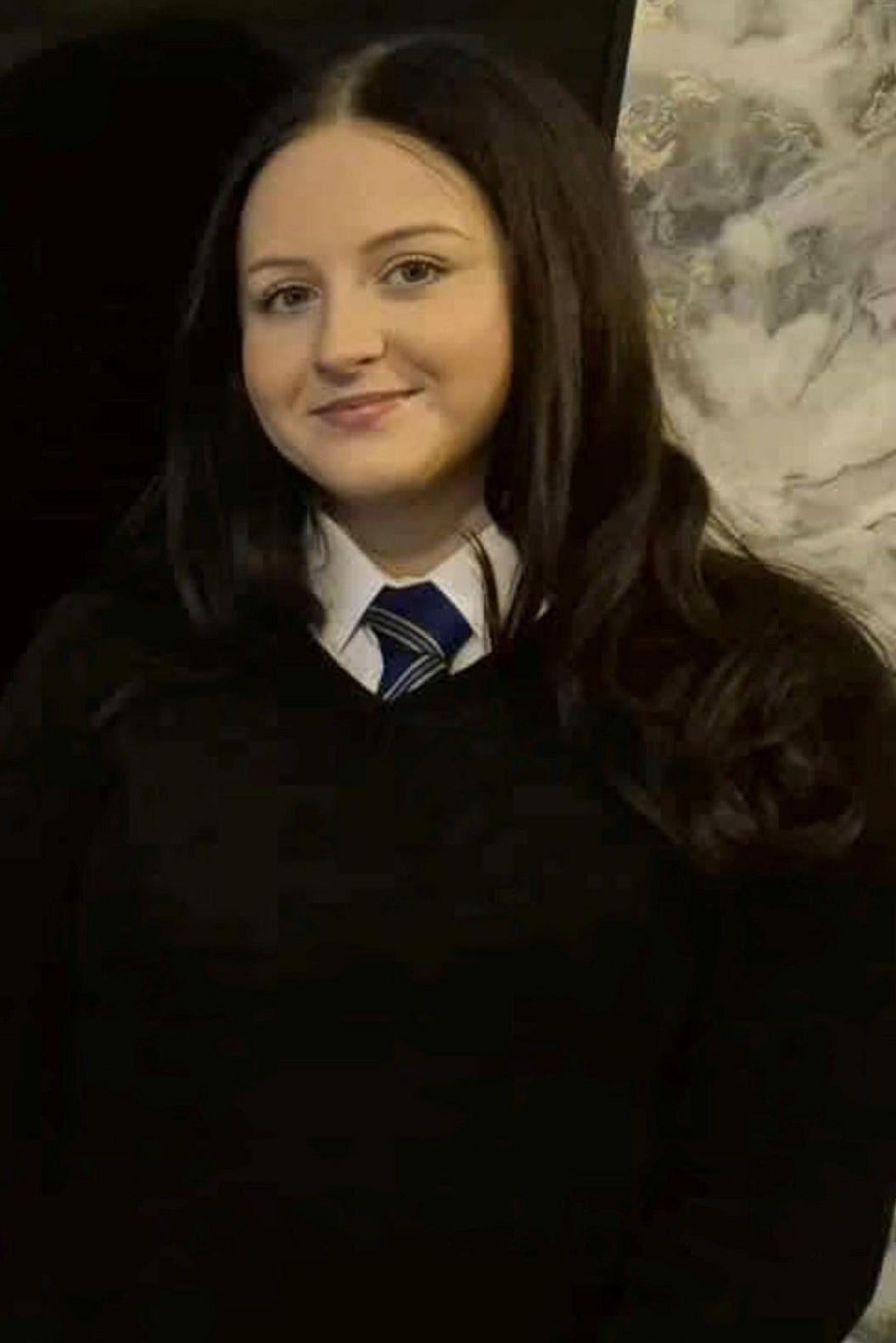 Lola smiles at the camera. she has brown hair and is wearing her school uniform