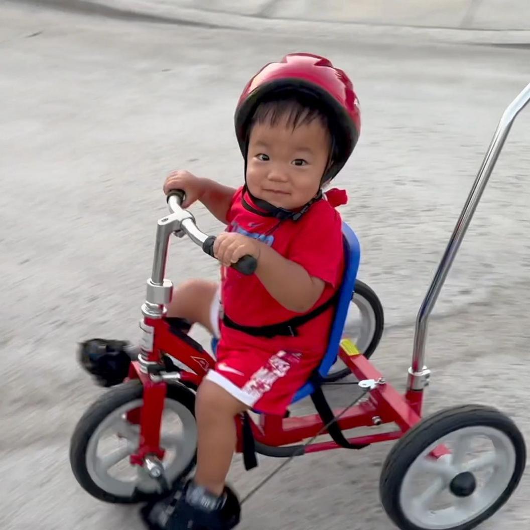 Oliver Chu rides a red tricycle. He is wearing a red helmet, red T-shirt and red and white shorts and is looking at the camera