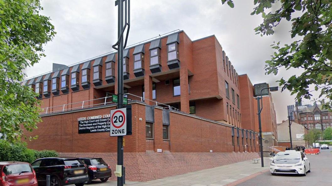 Exterior of Leeds Crown Court, a red brick building with windows jutting out on the top storey. A sign on the building says Leeds Combined Courts. Several cars are parked in front of the building.