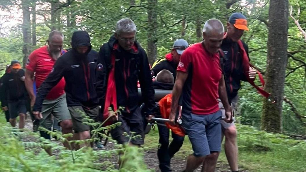 Mountain rescue team members carrying the man on a stretcher. They are wearing black and red uniform and walking along a gravel path in the woods.