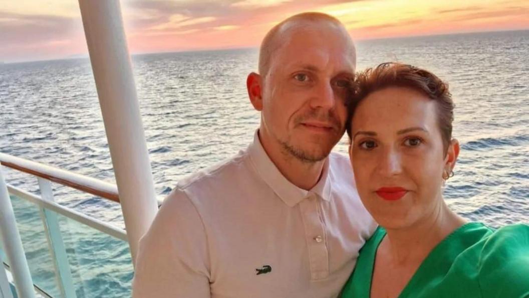 Rachel and her husband stand on the railing of a ship at sea as the sun sets in the background. She has short hard and a green dress and is leaning into his shoulder with her hair touching his cheek. He is a wearning a white lacoste t-shirt. 