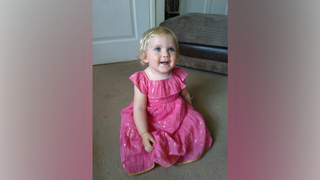 A young baby girl with blonde hair and blue eyes, wears a pink dress, as she stares up and smiles, in a living room