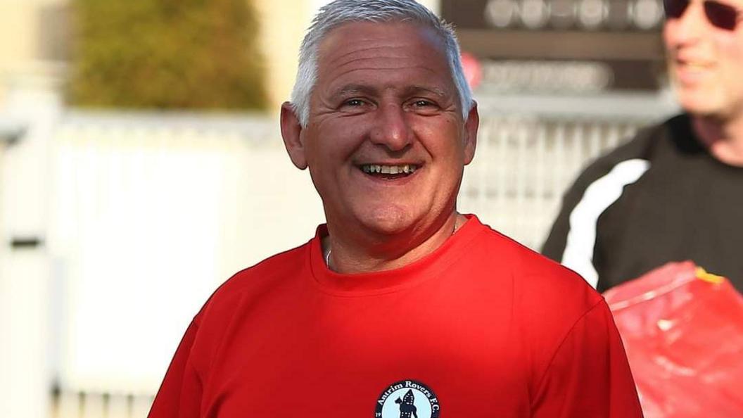 Mark Gourley wears a red top with an Antrim Rovers badge. He has grey hair and is shown here smiling.