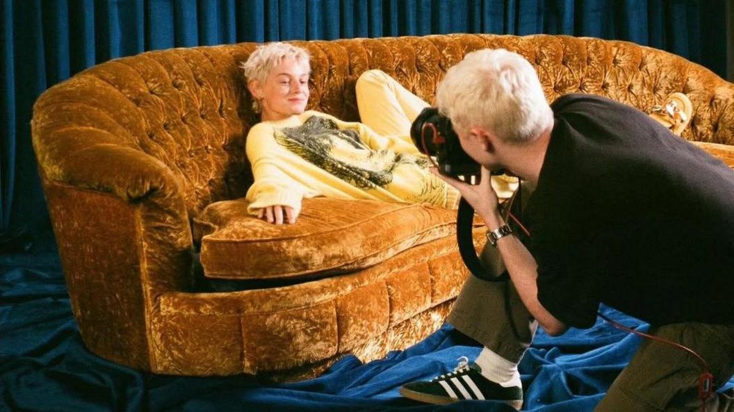 Emma Corrin lies on a orange velvet sofa, surrounded by blue curtains along the walls and floor. They have light blonde, short hair and are in a yellow jumper. Lloyd Wakefield is crouched in front of the sofa, with a camera in hand, taking a photo of Emma. He is wearing a black T-shirt and green trousers and has short blonde hair.