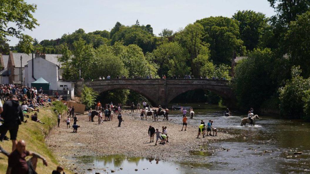 View downriver of people attending the Appleby Horse Fair. People are lining the banks of a river to watch riders taking their animals into the river. Others are watching from a bridge. The weather is sunny.