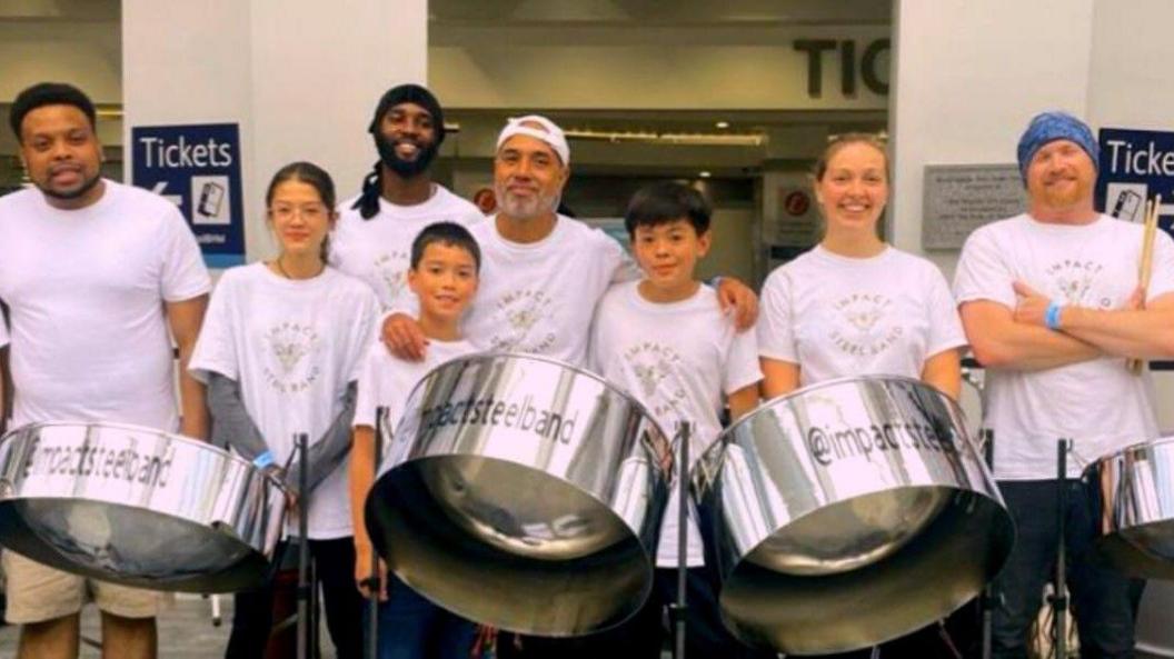 Eight people from the steel band. they are in white t-shirts that say "impact steel band". They are standing in front of three silver steel drums.
