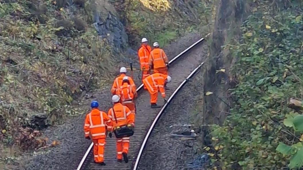 Seven men in orange high vis trousers and jackets, wearing helmets, walk on the train track. One is bent over to inspect a piece of the track, which is surrounded either side by a steep, rocky bank and plants