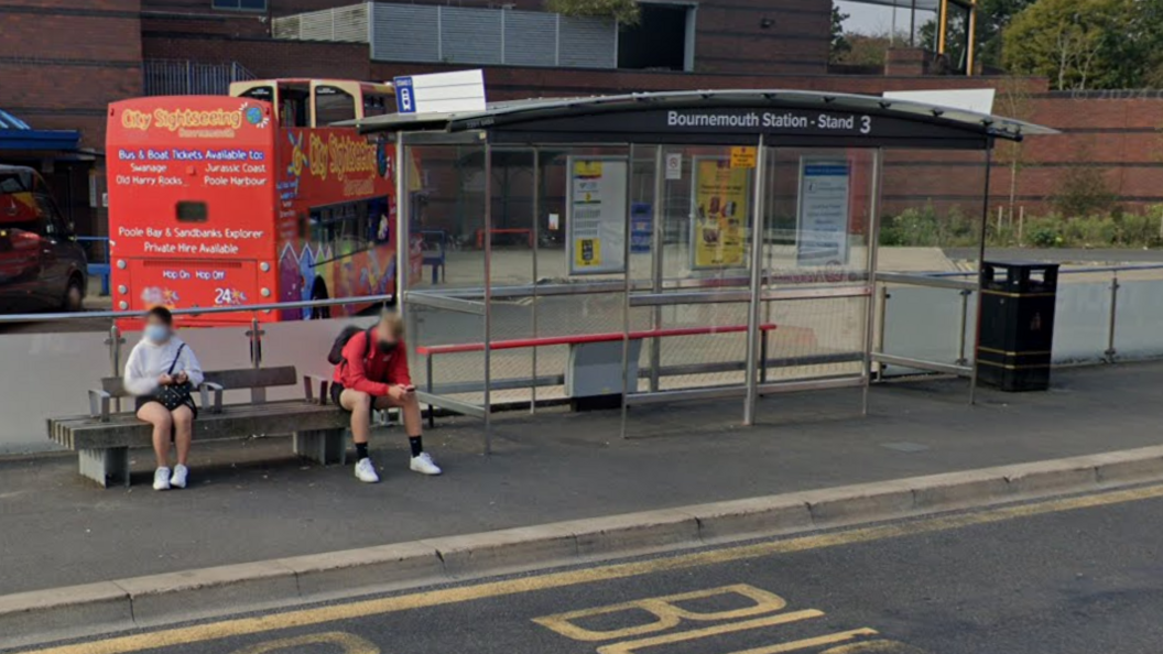 A man and a woman sitting on a bench next to a bus shelter. The bus shelter reads "Bournemouth Station - Stand 3". There is a red bus behind and a large brick building.