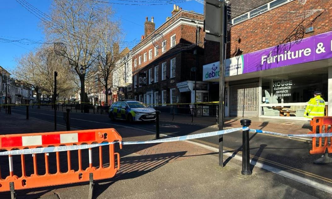 Orange hoardings and a police tape is strung between bollards on a high street with a police officer standing to the right and a police car parked on the high street outside a furniture shop