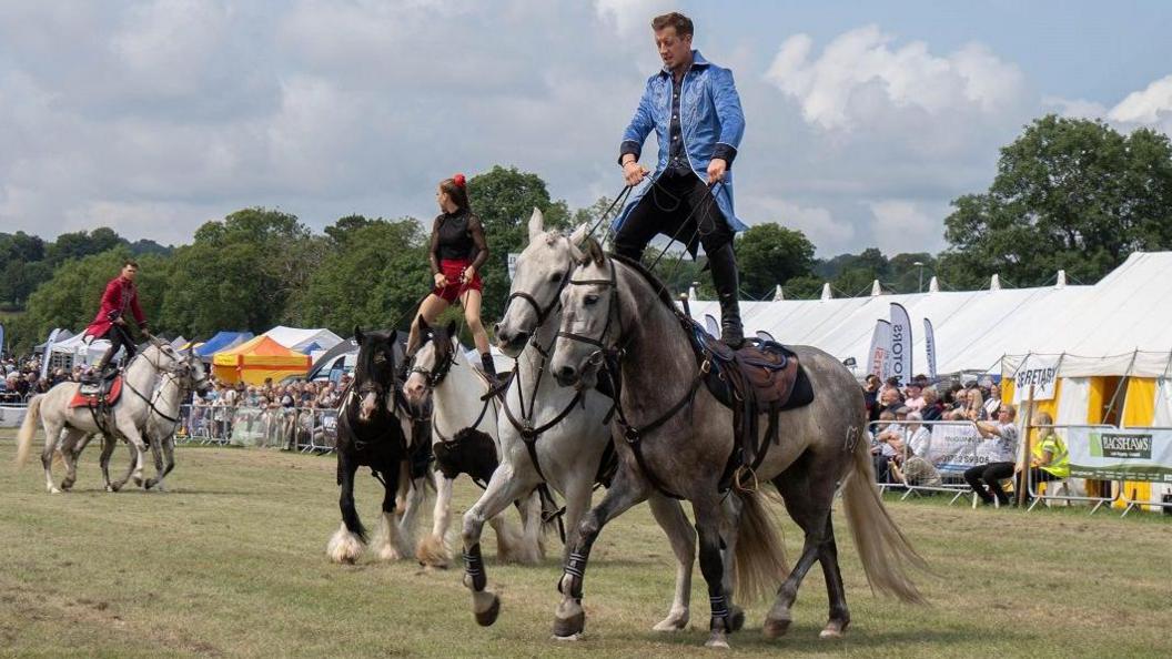 A man in a blue coat and black trousers stands with a foot on the back of two horses. Behind him, a woman in a black and red outfit does the same on two different horses. Behind them, an audience looks on, in front of white tents.