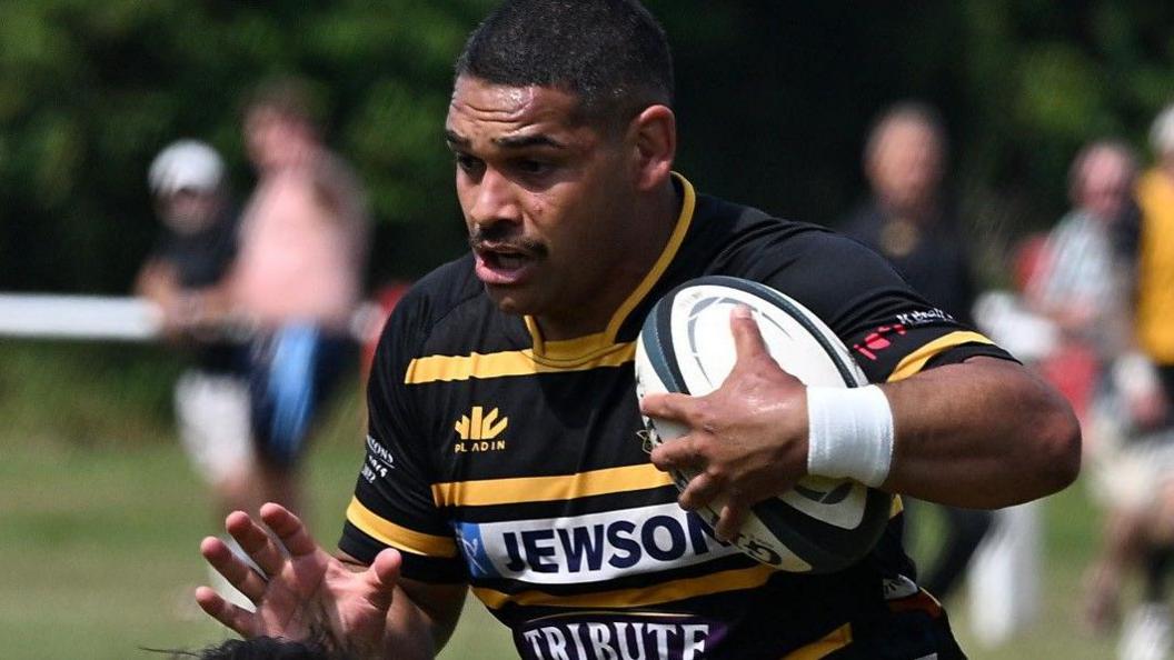 Sam Matavesi wears a black and yellow striped Cornwall shirt while in action against Kent, holding a rugby ball.