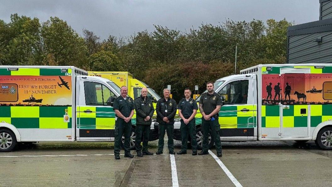 Five people, all wearing paramedic uniform, are stood in front of two ambulances which are facing each other. The ambulances have Remembrance Day designs on them.