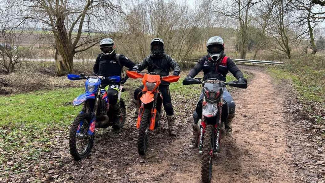 Three riders on off-road bikes side by side on a dirt track. They wear helmets and their faces are hidden