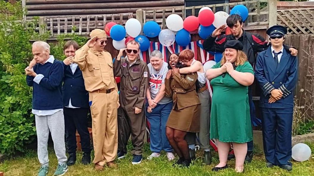A large group of people, dressed in costumes, standing by a fence, smiling, laughing, standing still and holding onto each other. There are balloons and decorations. 