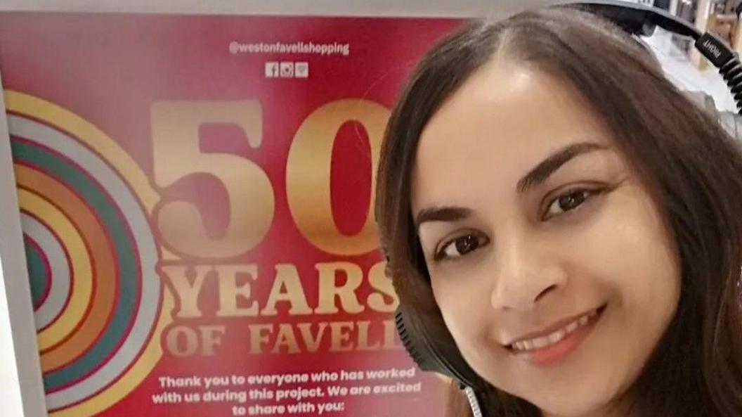 A close-up selfie of Subika Anwar-Khan, wearing headphones, in front of a red sign that reads '50 Years of Weston Favell'