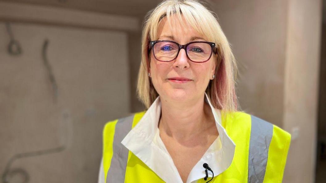 A woman with shoulder-length, straight blonde hair poses for a photo. She wears a white collared shirt under a high-vis gilet. There is a small black microphone pinned to the collar of her shirt. She has a pink lipstick on and wears glasses with black and dark pink frames. She is smiling softly at the camera. The background appears to be freshly-plastered walls, but it is in soft focus.