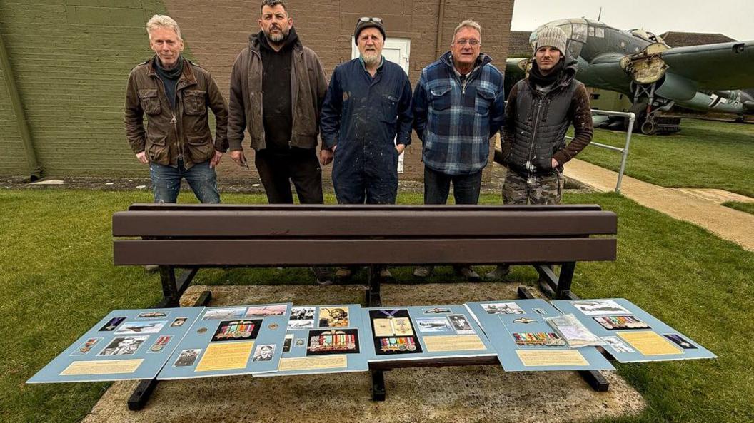 Five men standing behind a six sets of military medals which are laid on a wooden bench. 