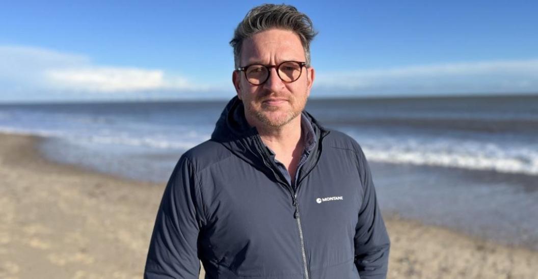 Man with glasses looking directly at the camera while standing on a beach. he is wearing a blue jacket.