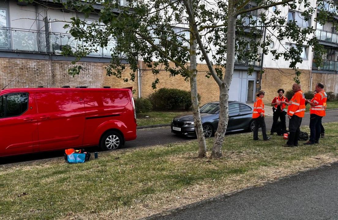 People, wearing orange jackets, stand on a grass verge in front of a block of flats  in Greenwich 