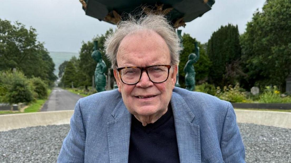 An elderly man with grey hair and black square glasses and a grey blazer on. He is standing in front of some grey stones and blue statues and what looks like a public garden with trees and grass. 
