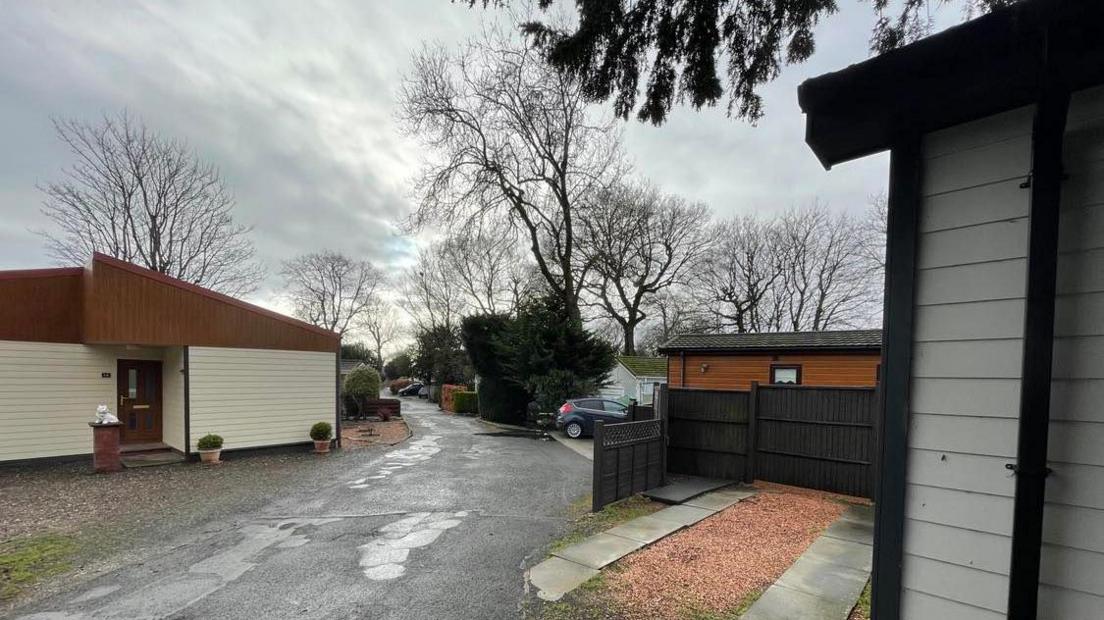 A view of some of the park homes - one has white walls with brown door and roof, with a bin sitting in front of it. Another has brown walls and a dark roof, with a car parked next to it. Other properties can be seen in the distance, as can other cars.