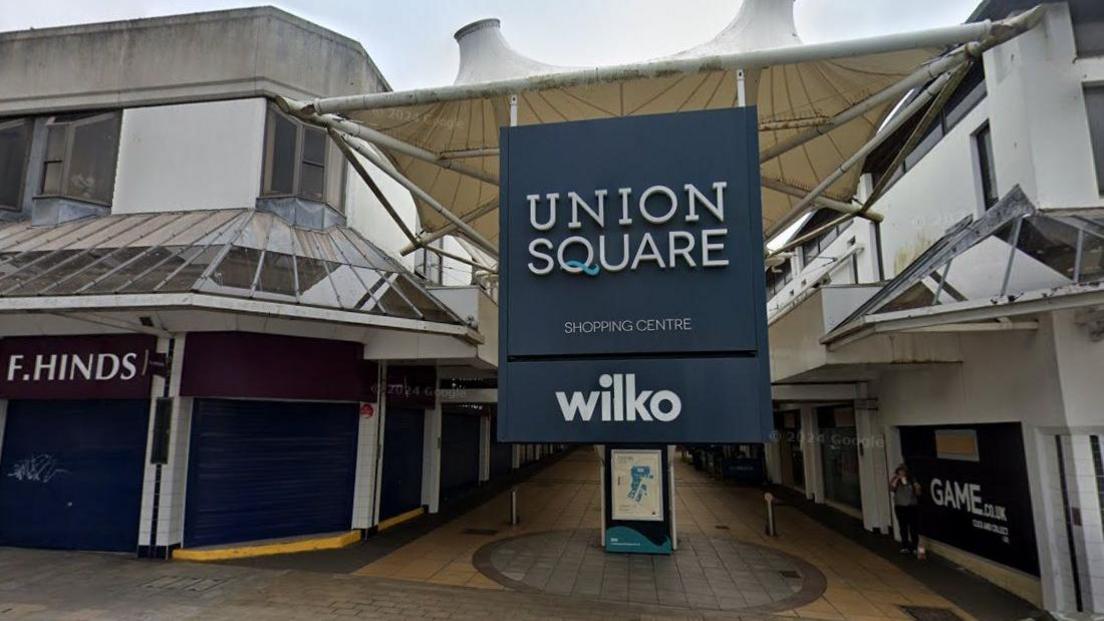 The outside of the Union Square shopping centre in Torquay. There is a large square navy blue sign that reads Union Square with shops either side.