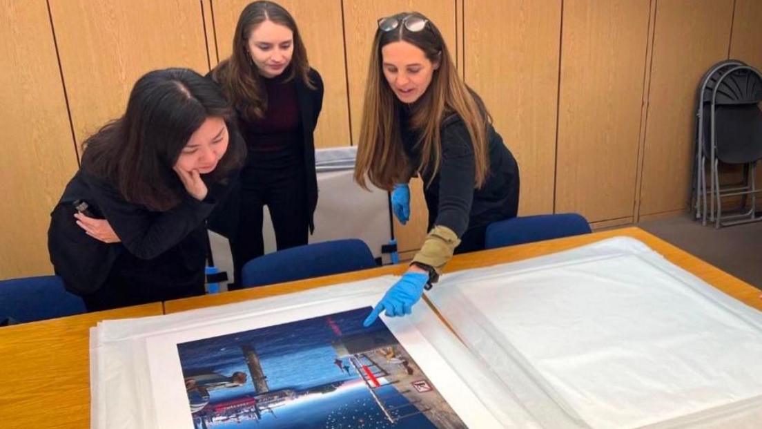 Three women dressed in black looking at artwork on a table, Ms Allchurch is wearing blue latex gloves