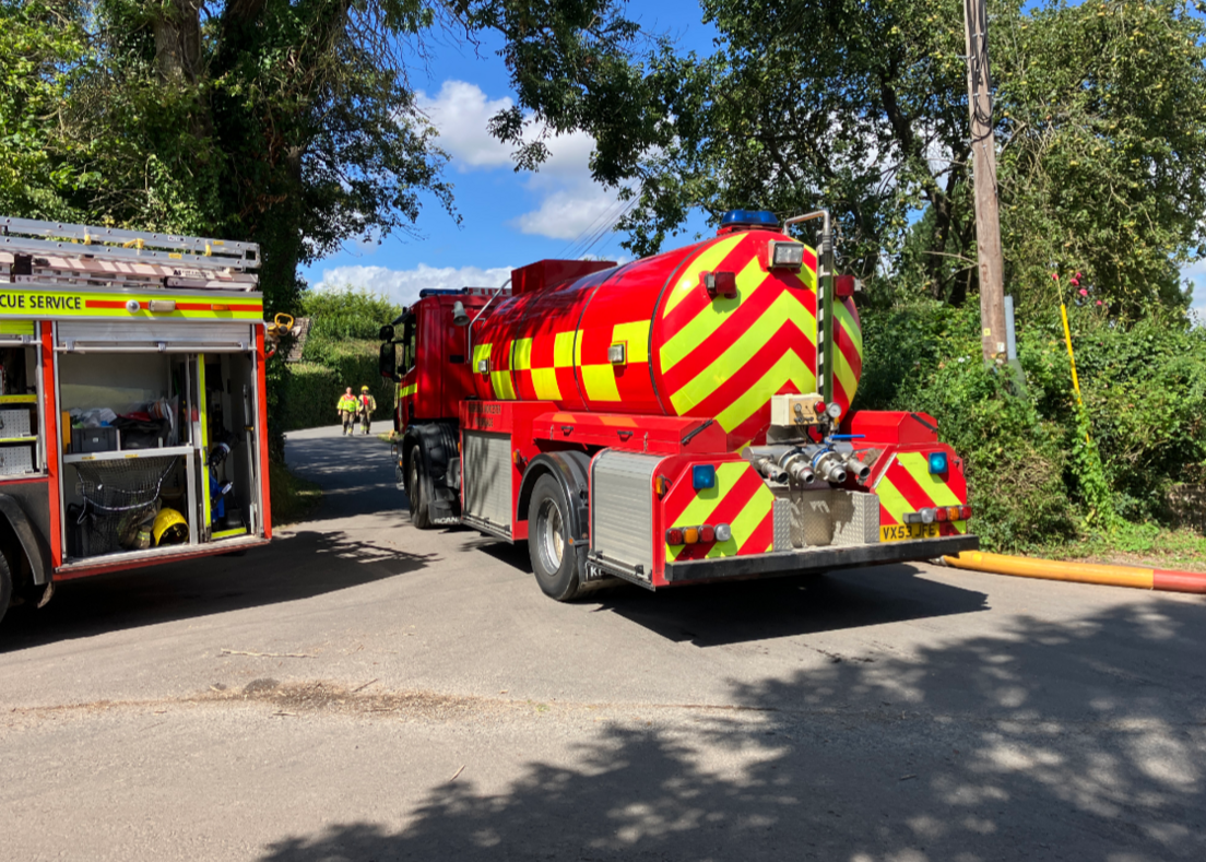 A large fire service water tanker to the right, fire engine to the left and firefighters in the distance