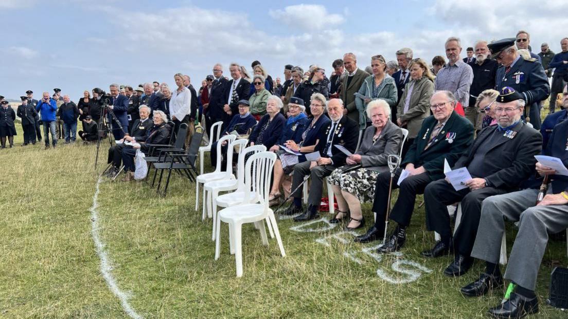 A group of elderly people on chairs listening to the Last Post being played 