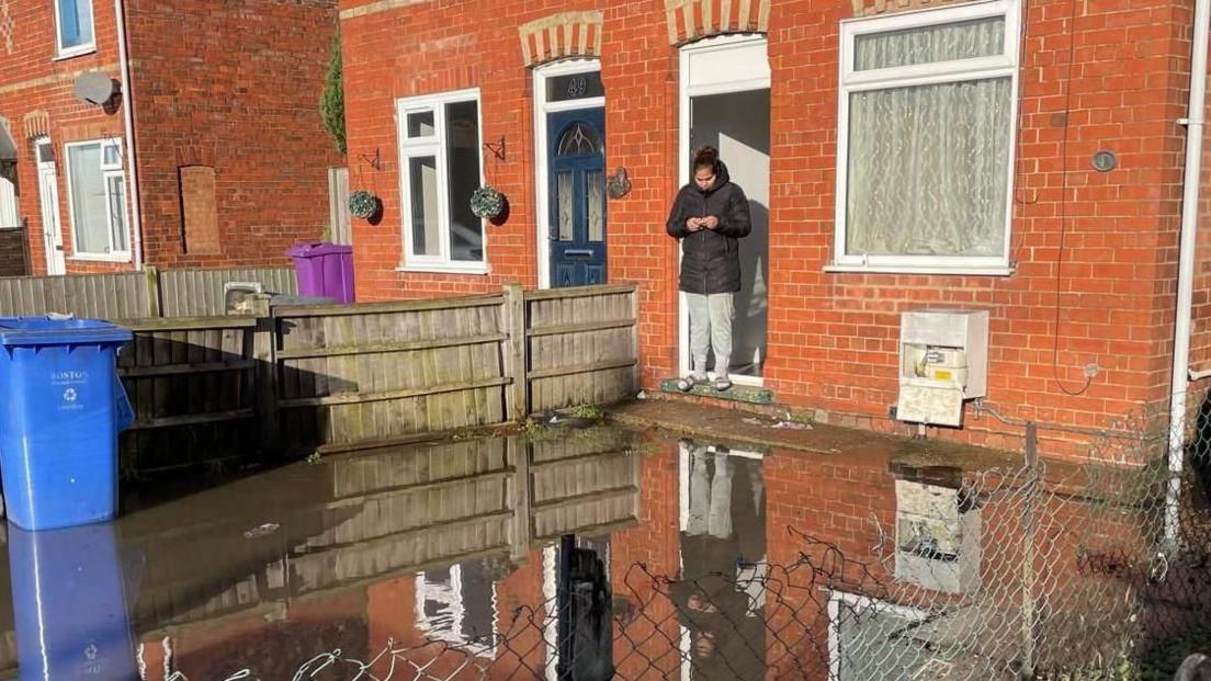 A flooded garden with a woman standing in a doorway cut off by water taking pictures of the garden.